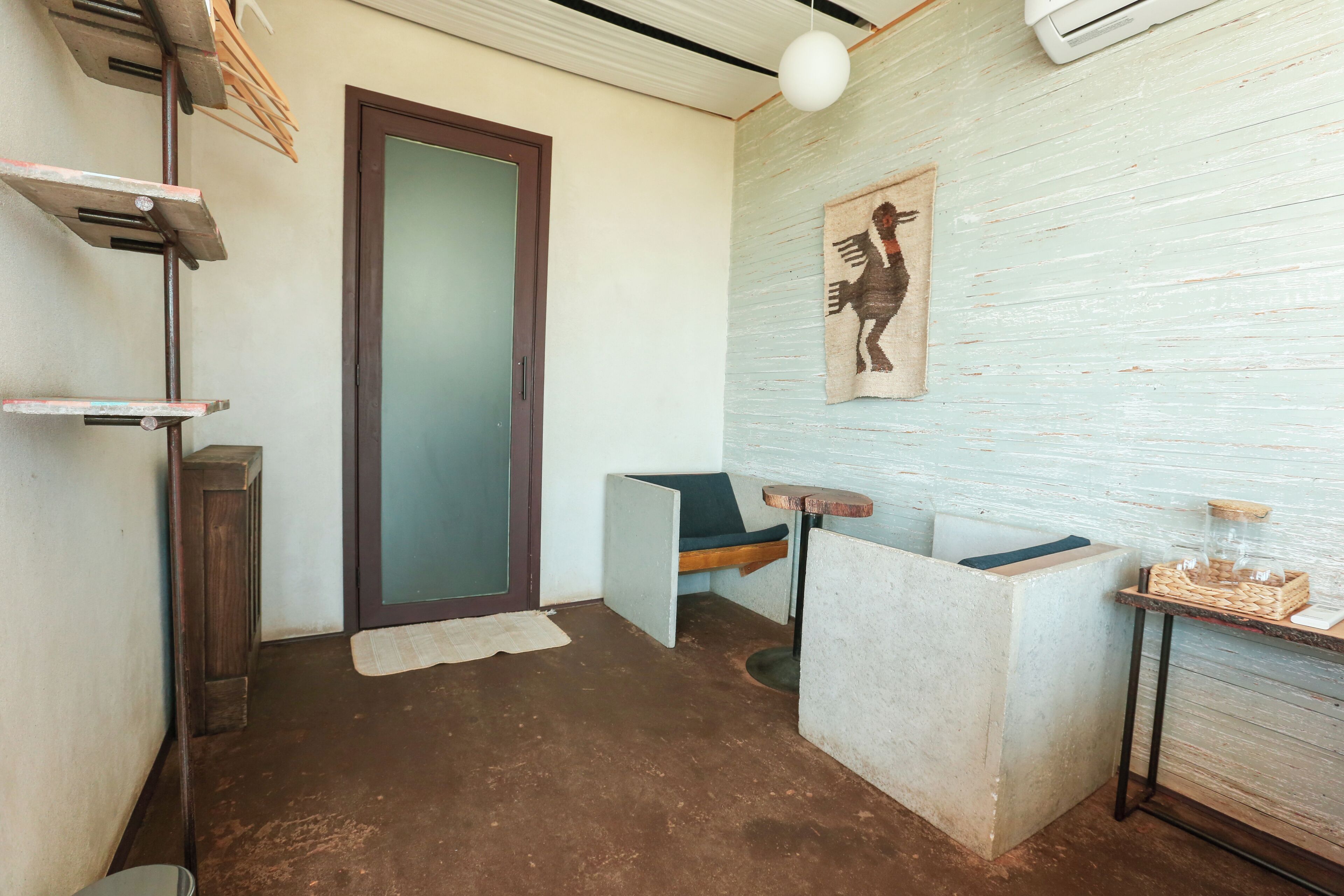 A compact room with a concrete bench, a small table with a basket, a wall-mounted shelf, and a door.