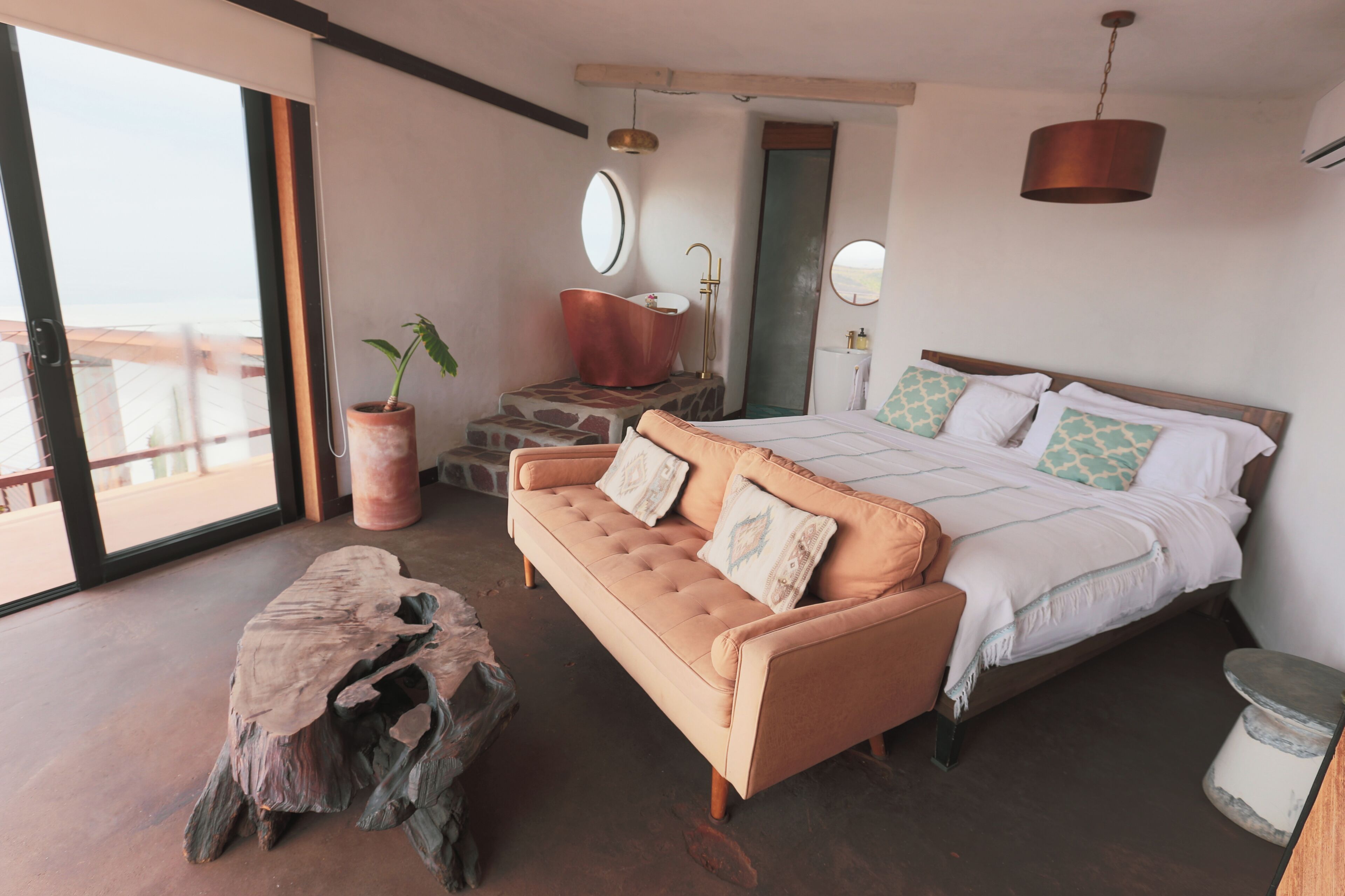Ein Schlafzimmer mit einem großen Bett, einer Couch, einem hölzernen Couchtisch, einer Topfpflanze und Blick durch ein großes Fenster.