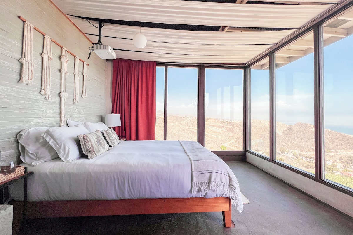 A bedroom with a large bed, a red curtain, a wall-mounted projector, and a view of the landscape through large windows.