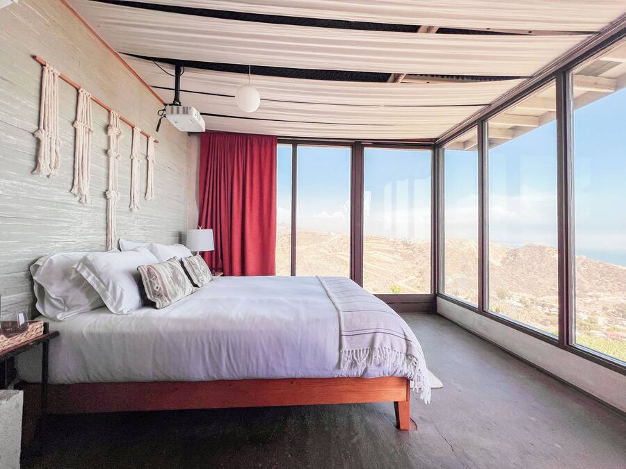 A bedroom with a large bed, a red curtain, a wall-mounted projector, and a view of the landscape through large windows.