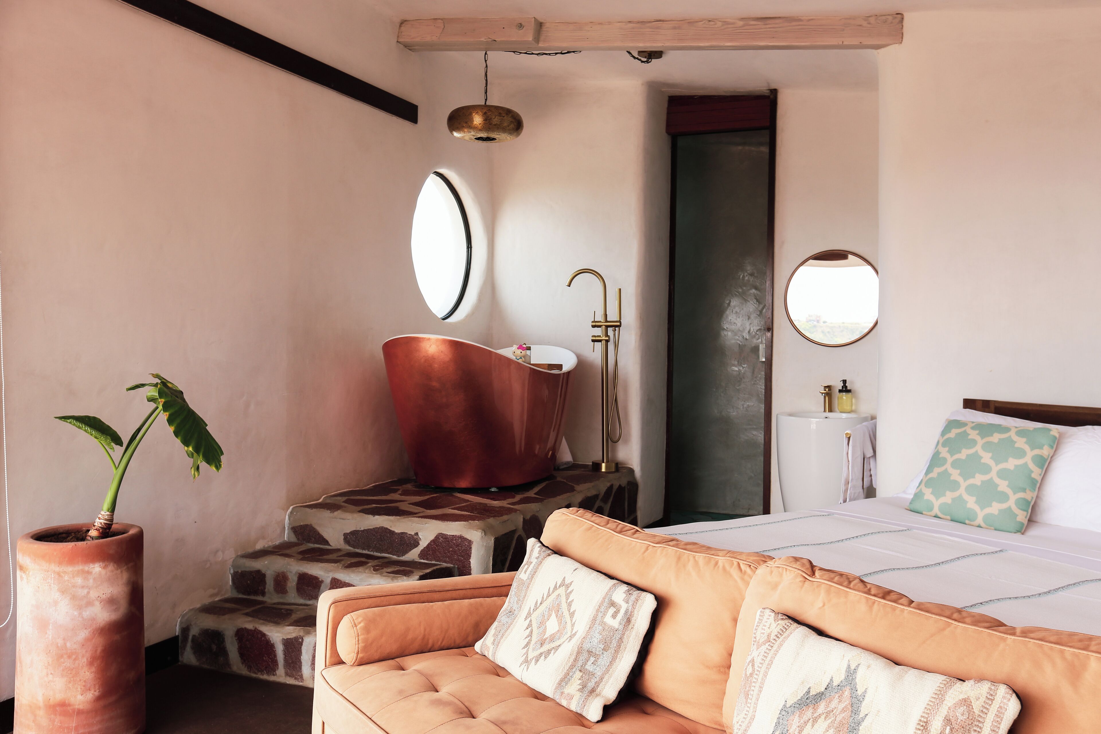 Une chambre à coucher avec un lit, un canapé, une grande plante en pot, une baignoire en cuivre et un grand miroir rond accroché au mur.