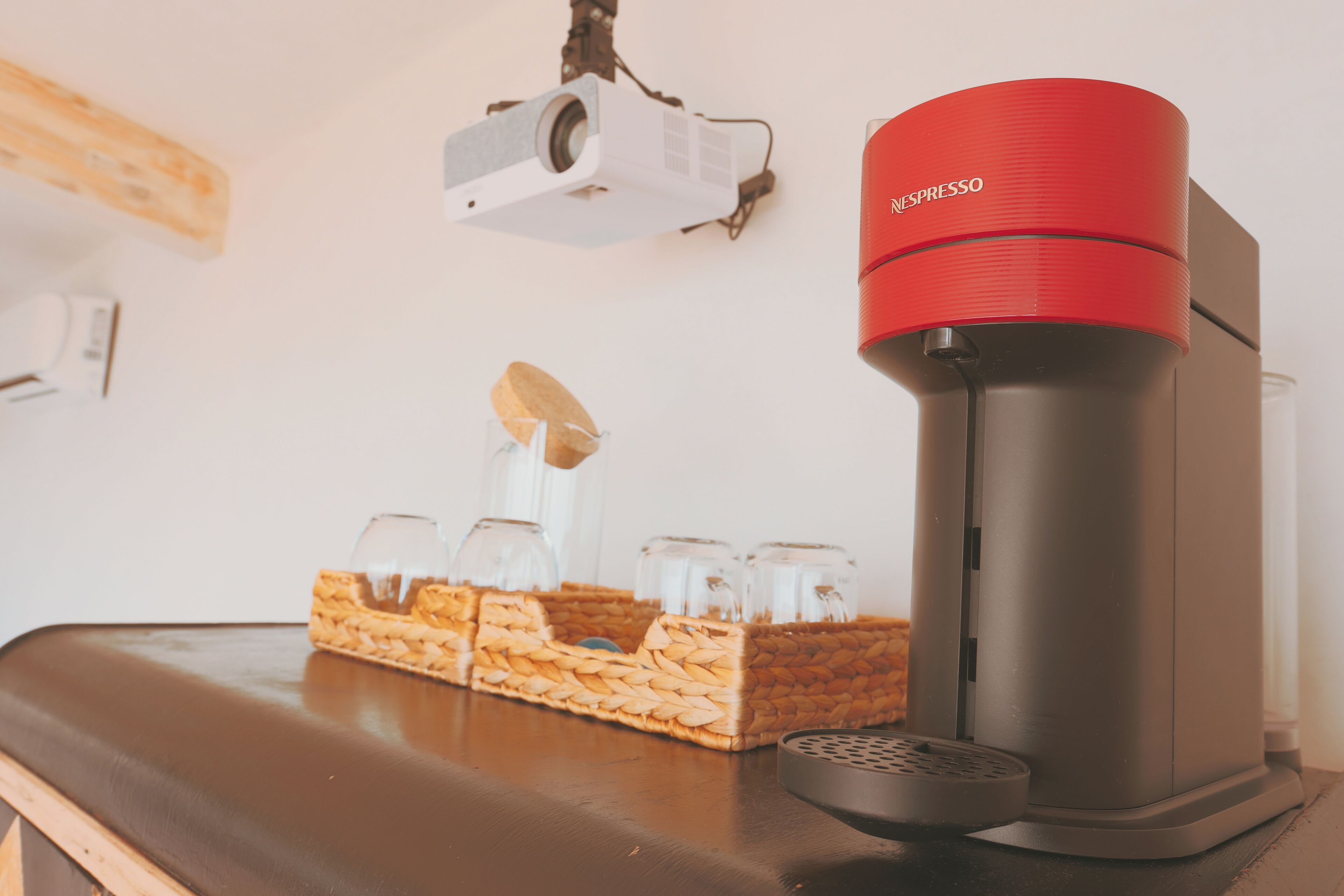 Una macchina da caffè con un piano superiore rosso, un proiettore e un cesto con bicchieri su un tavolo.