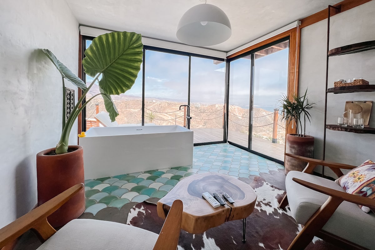 A modern bathroom with a bathtub, large windows, and a view of a mountainous landscape.
