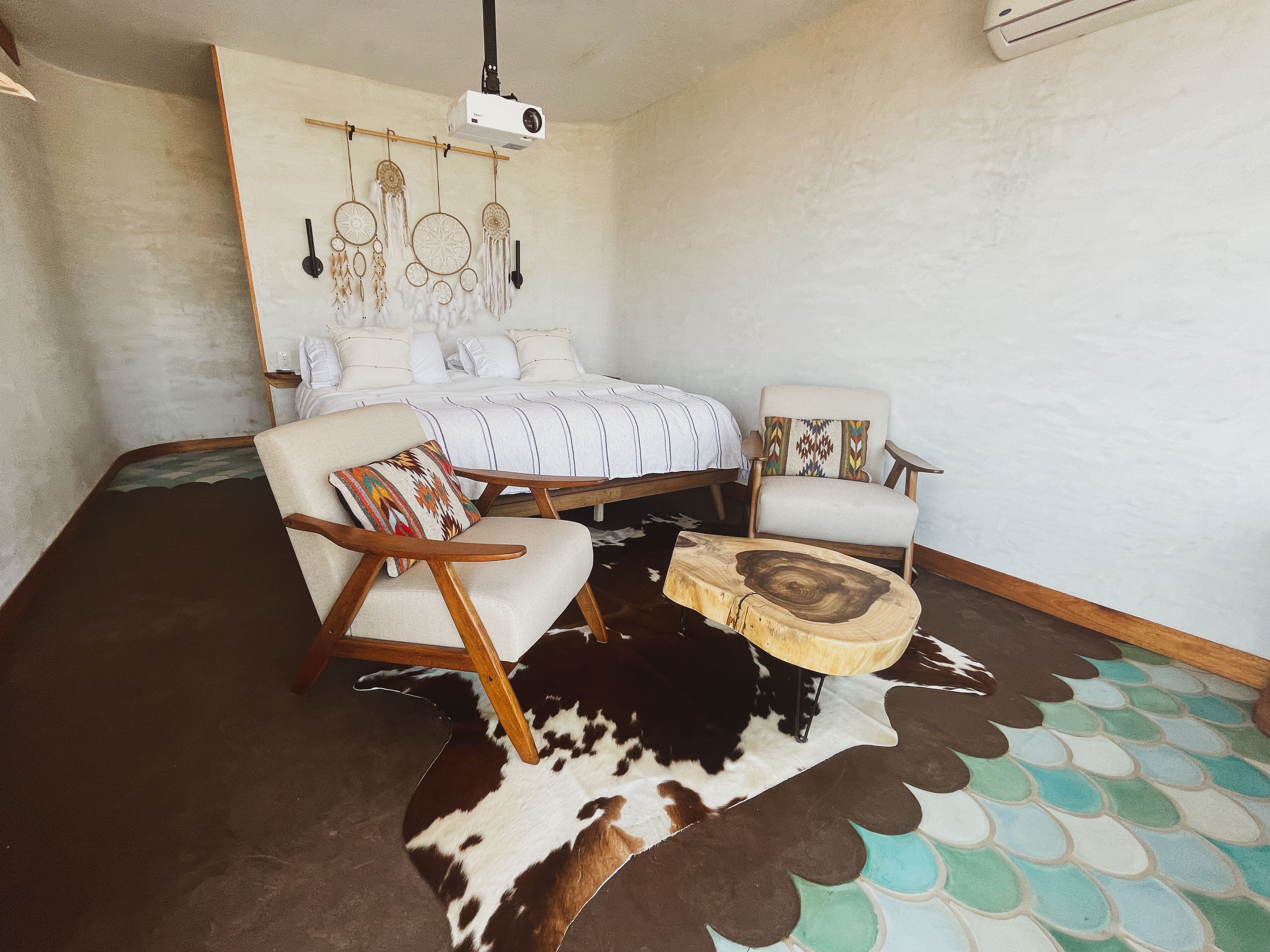 Un dormitorio con una cama, dos sillas, un proyector y una alfombra de cuero de vaca.