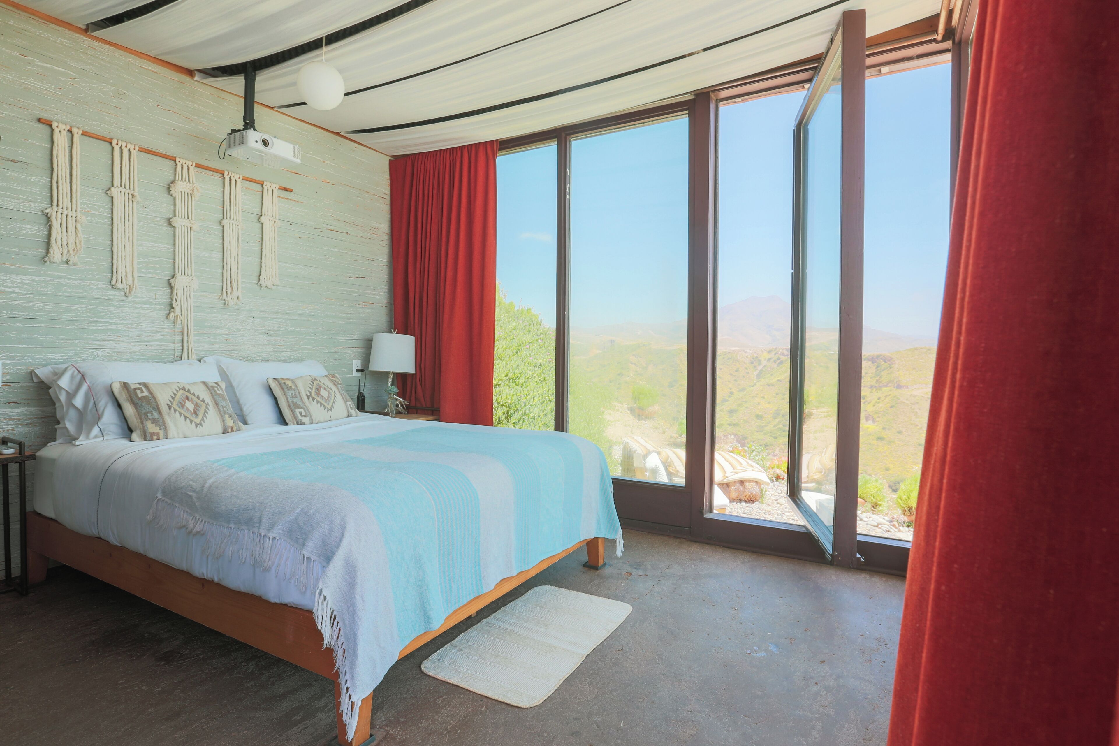A bedroom with a large bed, a nightstand, a lamp, and a view of the outdoors through large windows.