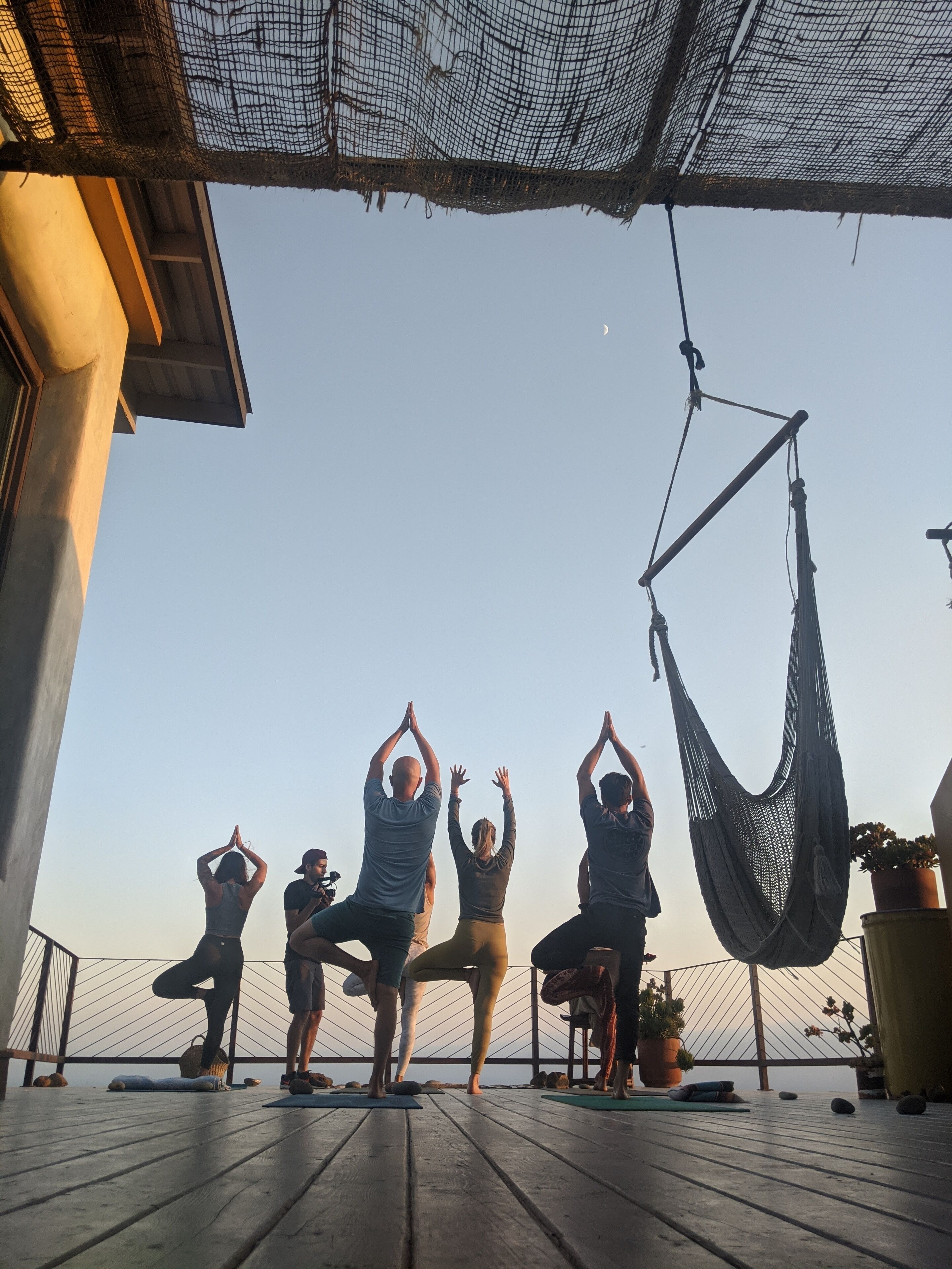 A group of people performing yoga on a wooden deck with a hammock and potted plants.