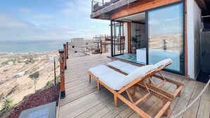 A modern outdoor deck with a wooden lounge chair and a glass-enclosed area with a view of the ocean. - Native Residence (Playas de Rosarito)