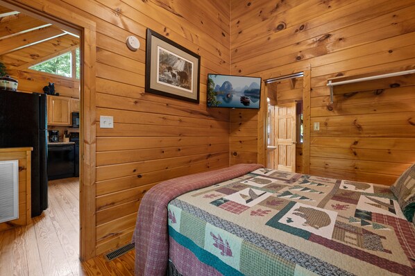 A wooden cabin interior with a bed, a kitchen area, and a mounted television.