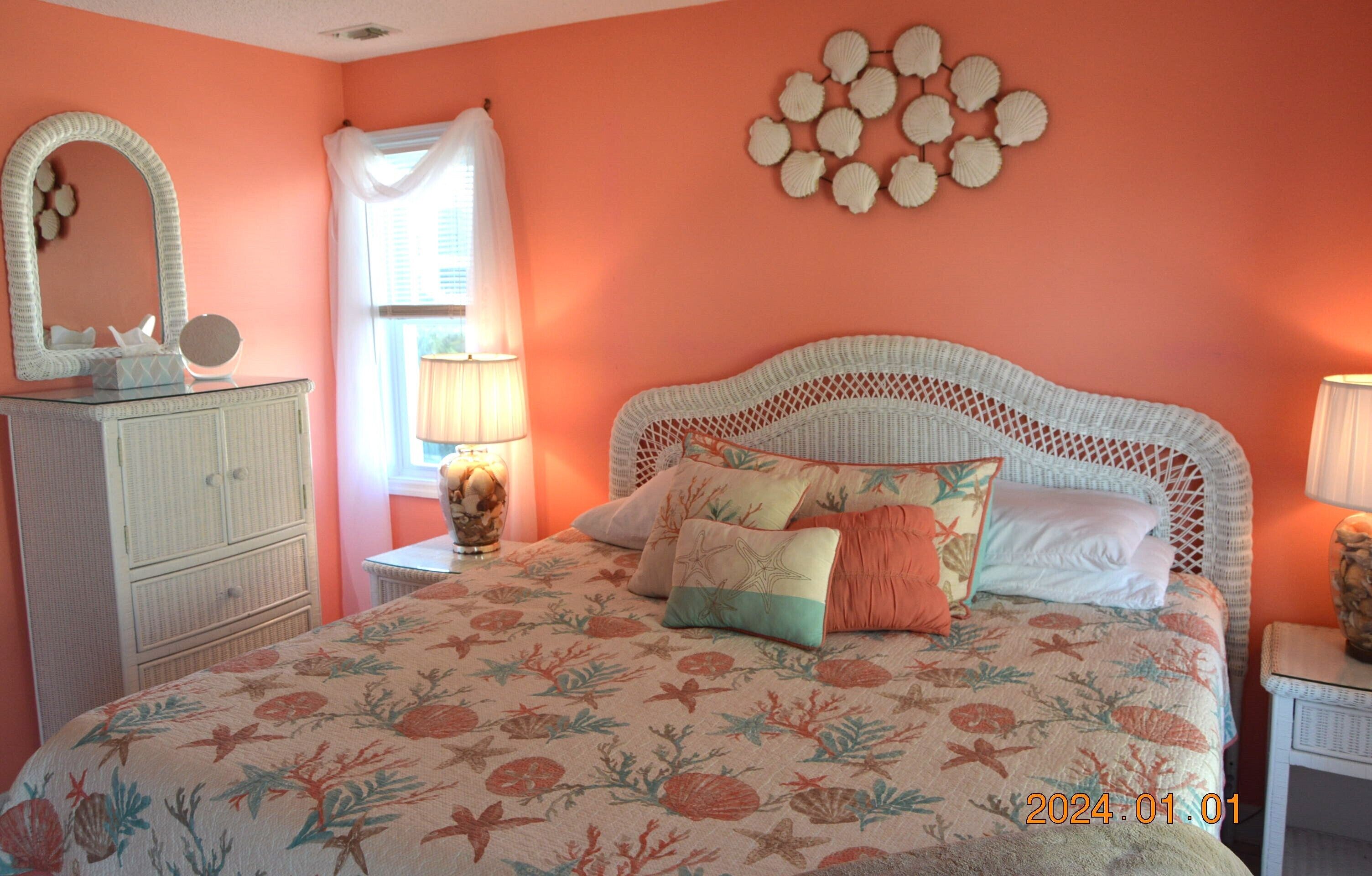A bedroom with a floral bedspread, a white wicker headboard, a dresser, and a wall decoration of shells.