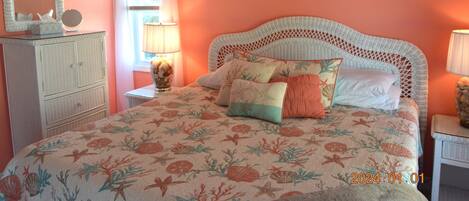 A bedroom with a floral bedspread, a white wicker headboard, a dresser, and a wall decoration of shells.