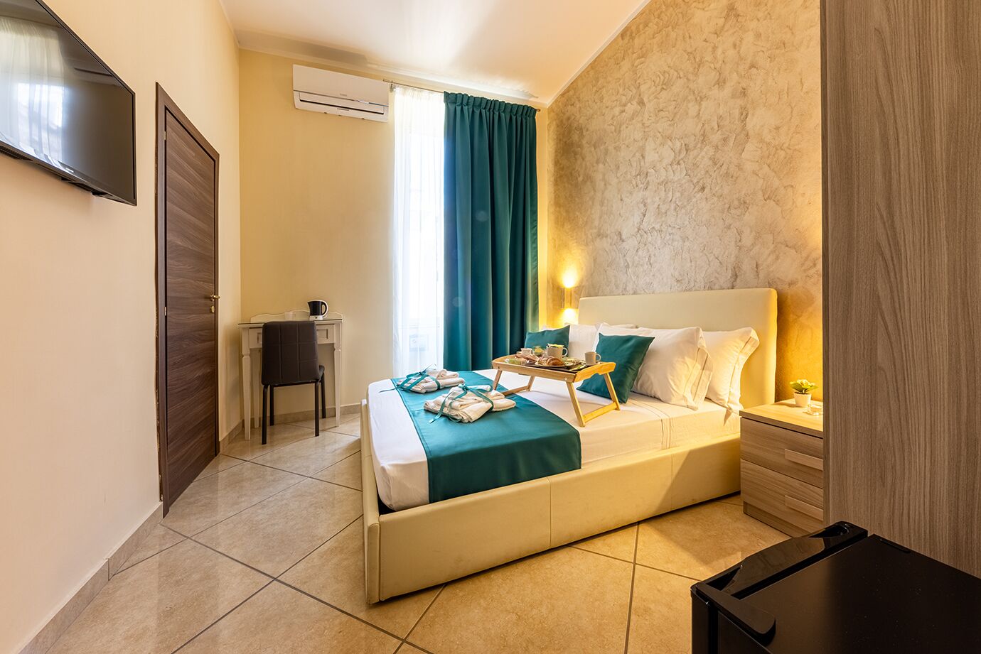 A hotel room with a bed, a desk, a chair, a TV, and an air conditioning unit.
