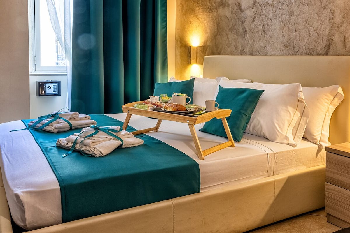 A hotel room with a bed, a bedside table with a tray of breakfast, a window with curtains, and a wall-mounted light.