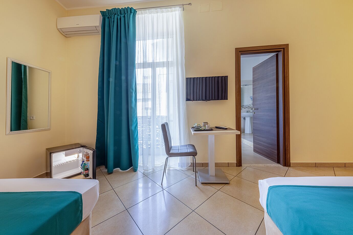 A hotel room with two beds, a desk, a chair, a TV, and a bathroom visible through an open door.