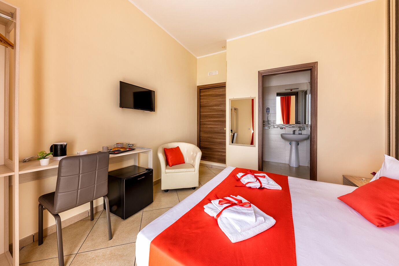 A hotel room with a bed, a desk, a chair, a TV, a mirror, and a bathroom.