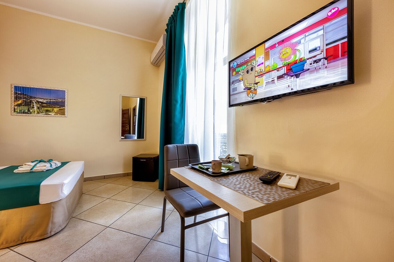 A hotel room with a bed, a desk, a chair, a TV, a window with curtains, a trash bin, and a small table with a tray.