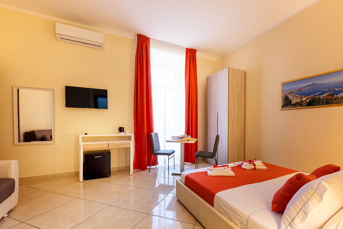 A hotel room with a bed, a desk, a chair, a TV, and a window with red curtains.
