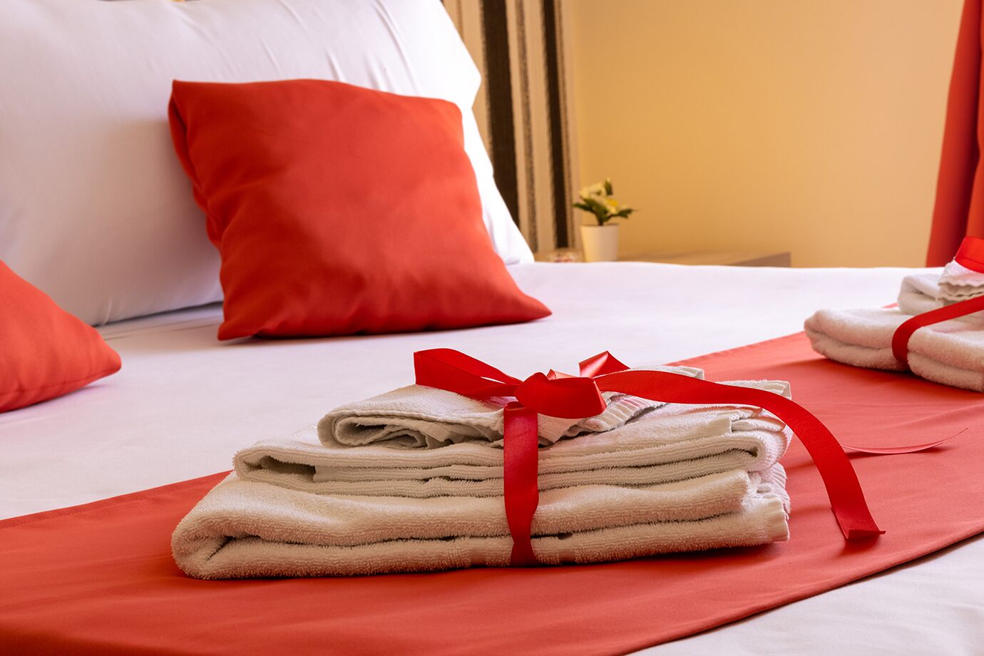 A neatly made bed with red pillows and a red blanket, a stack of folded towels with a red ribbon, and a small potted plant on a bedside table.