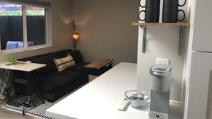 A compact kitchen with a white countertop, a coffee maker, and a kettle. There is a black sofa with a patterned pillow and a small table with a green cup and a book. A window with blinds is visible, and a wall-mounted shelf holds two black mugs. - Artfully Curated Loft-Like Studio (Culver City)