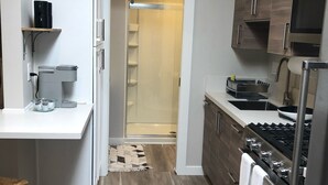 A modern kitchen with a built-in stove, microwave, and oven. There is a small island with a sink and a coffee maker. The kitchen has wooden flooring and a glass shower door leading to a bathroom. - Artfully Curated Loft-Like Studio (Culver City)