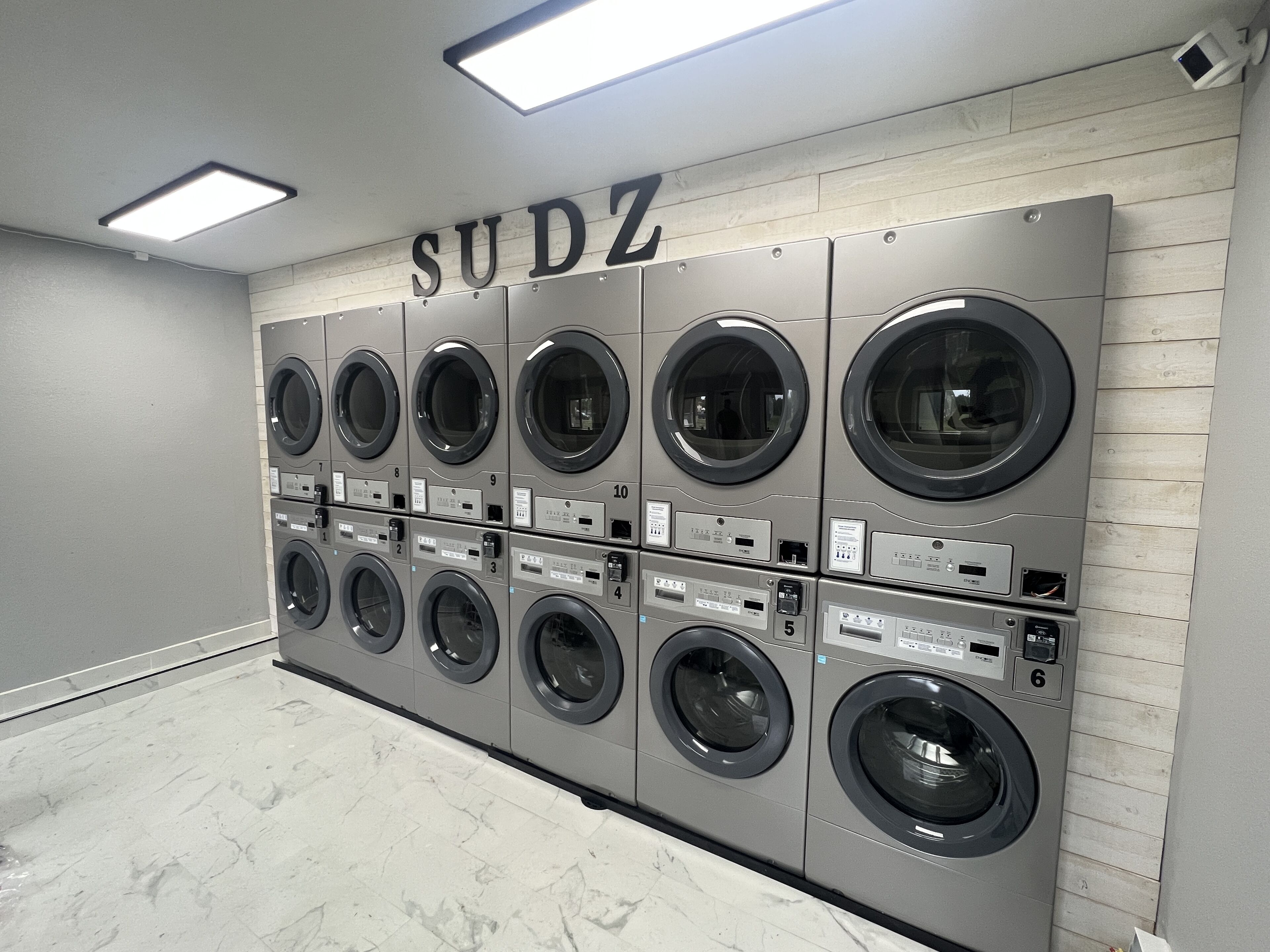 Une rangée de six machines à laver fixées au mur dans une buanderie, avec une enseigne au-dessus indiquant « SUDZ ».