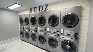 Une rangée de six machines à laver fixées au mur dans une buanderie, avec une enseigne au-dessus indiquant « SUDZ ».