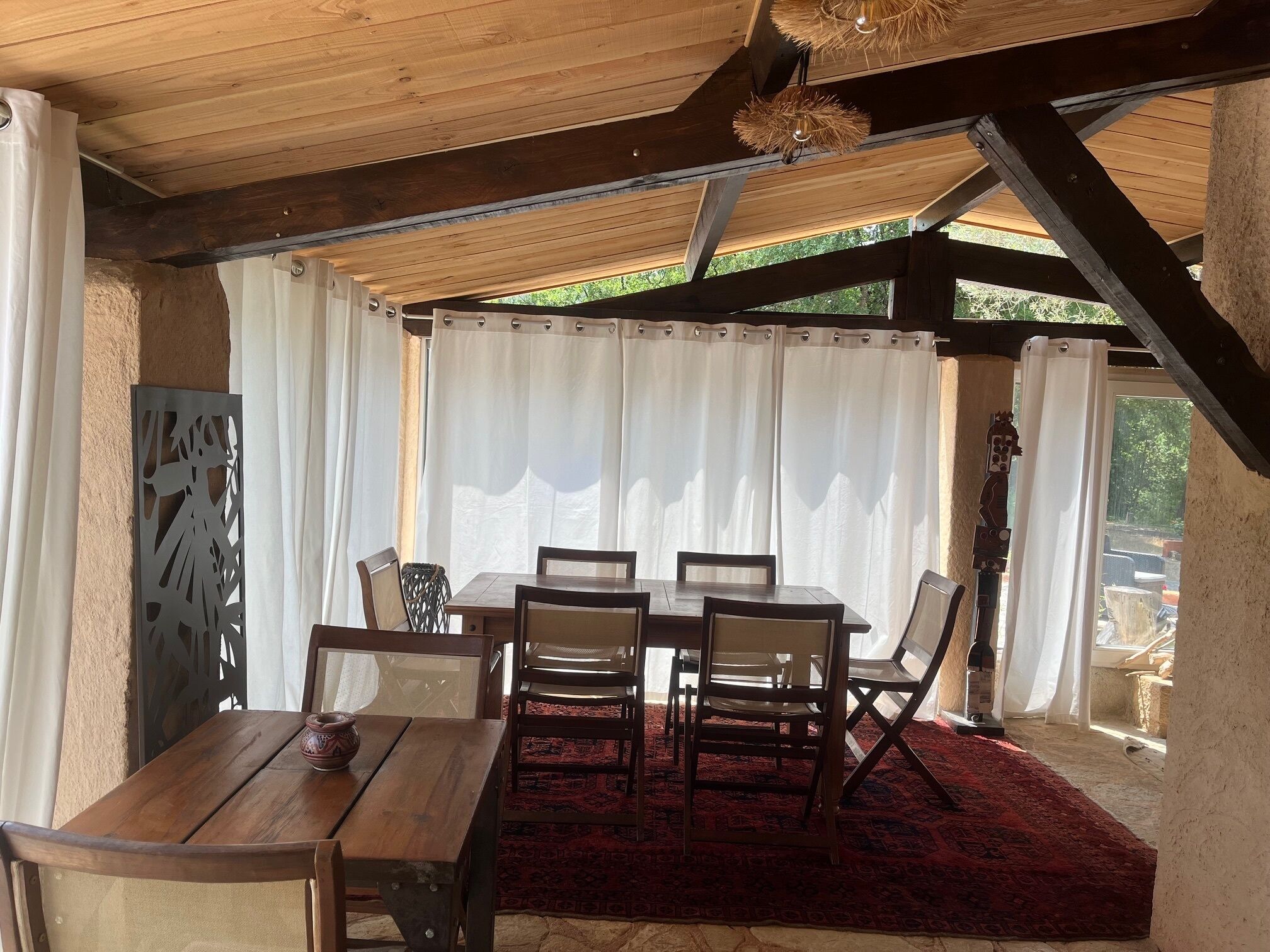 Un área de comedor al aire libre techada con una mesa y sillas de madera, cortinas blancas y un tapete rojo.