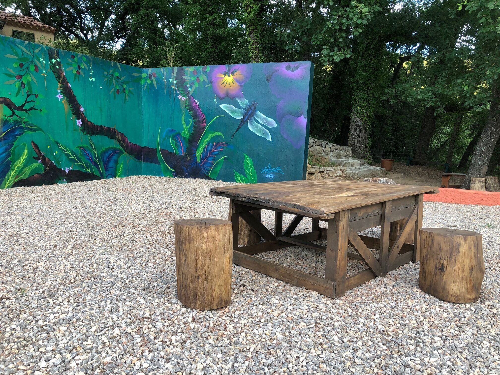 Uma mesa de madeira com bancos em uma área de cascalho, com um mural colorido ao fundo.
