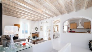 A modern interior with a glass table, white walls, wooden ceiling beams, and a kitchen area with wooden cabinets.