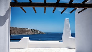 A view from a white-walled room with a tiled floor, looking out to a clear blue sea and a rocky hillside.