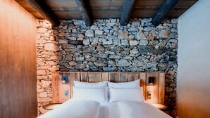 A bedroom with a stone wall headboard, wooden beams, and two bedside lamps. - Hotel Les Pardines 1819 (Encamp)
