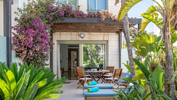 A patio area with a dining table and chairs, a sofa with cushions, and lush tropical plants.