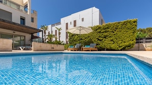 A swimming pool with tiled blue edges, a white building with a balcony, and a green hedge.