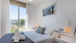 A hotel room with a large bed, a nightstand with a lamp, a painting on the wall, and a view of the outdoors.