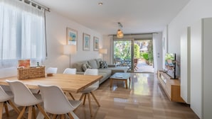 A modern living room with a wooden dining table, white chairs, a sofa, and a TV on a wooden stand.