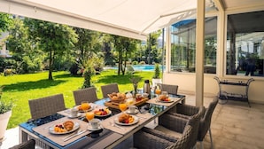 An outdoor dining area with a table set for breakfast, surrounded by wicker chairs, overlooking a pool and lush garden. - Villa Guidi 10, Emma Villas (Cervia)