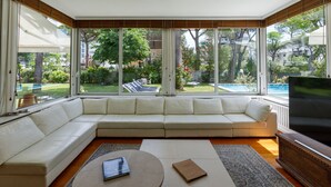 A modern living room with a large sectional sofa, a flat-screen TV, and a view of a swimming pool and greenery. - Villa Guidi 10, Emma Villas (Cervia)