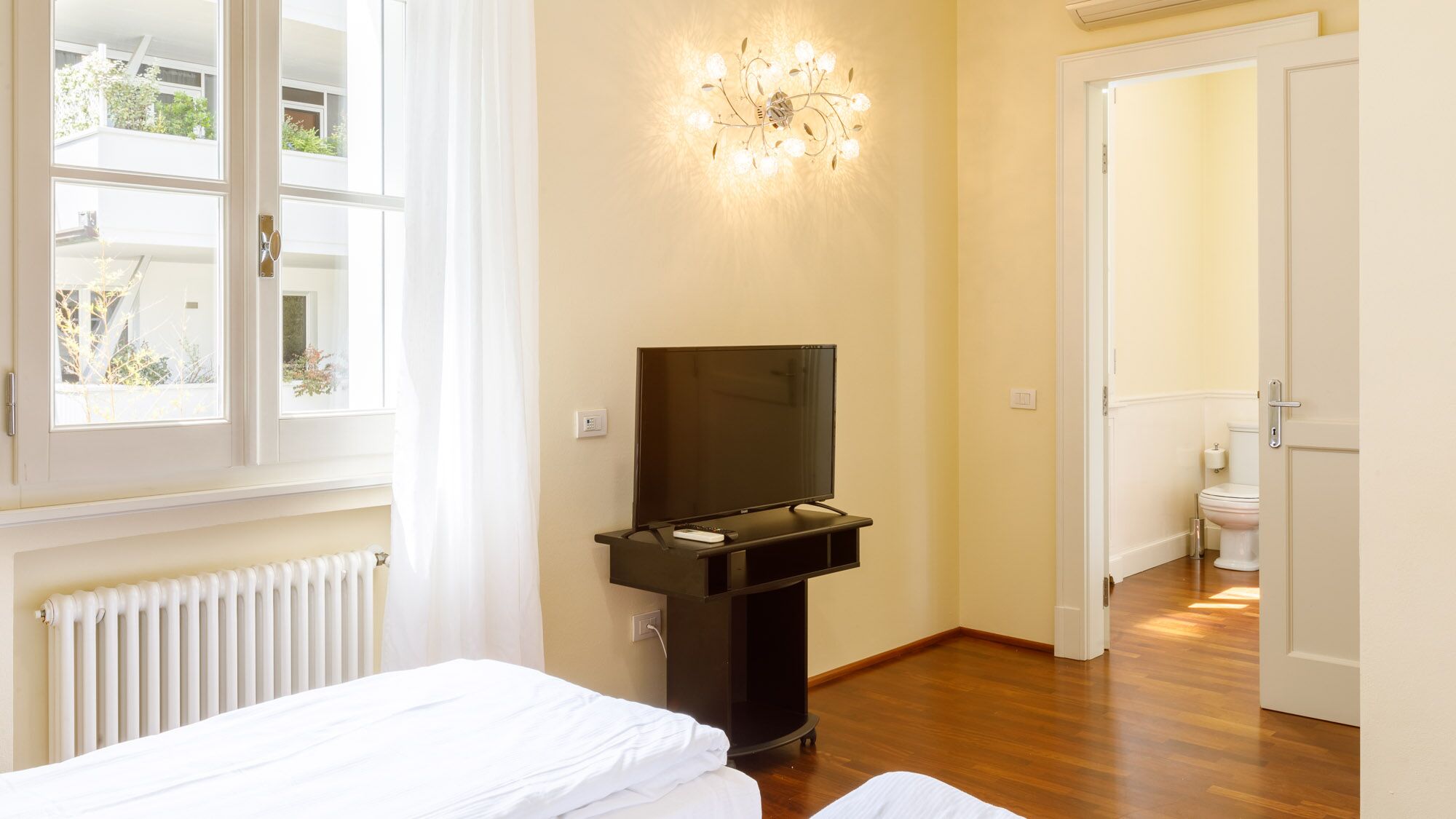A hotel room with a bed, a television on a stand, a radiator, and a window with curtains.