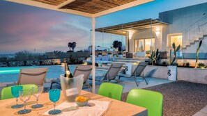 A poolside dining area with outdoor seating, a table with a bottle of champagne, and a view of the pool and a modern building.