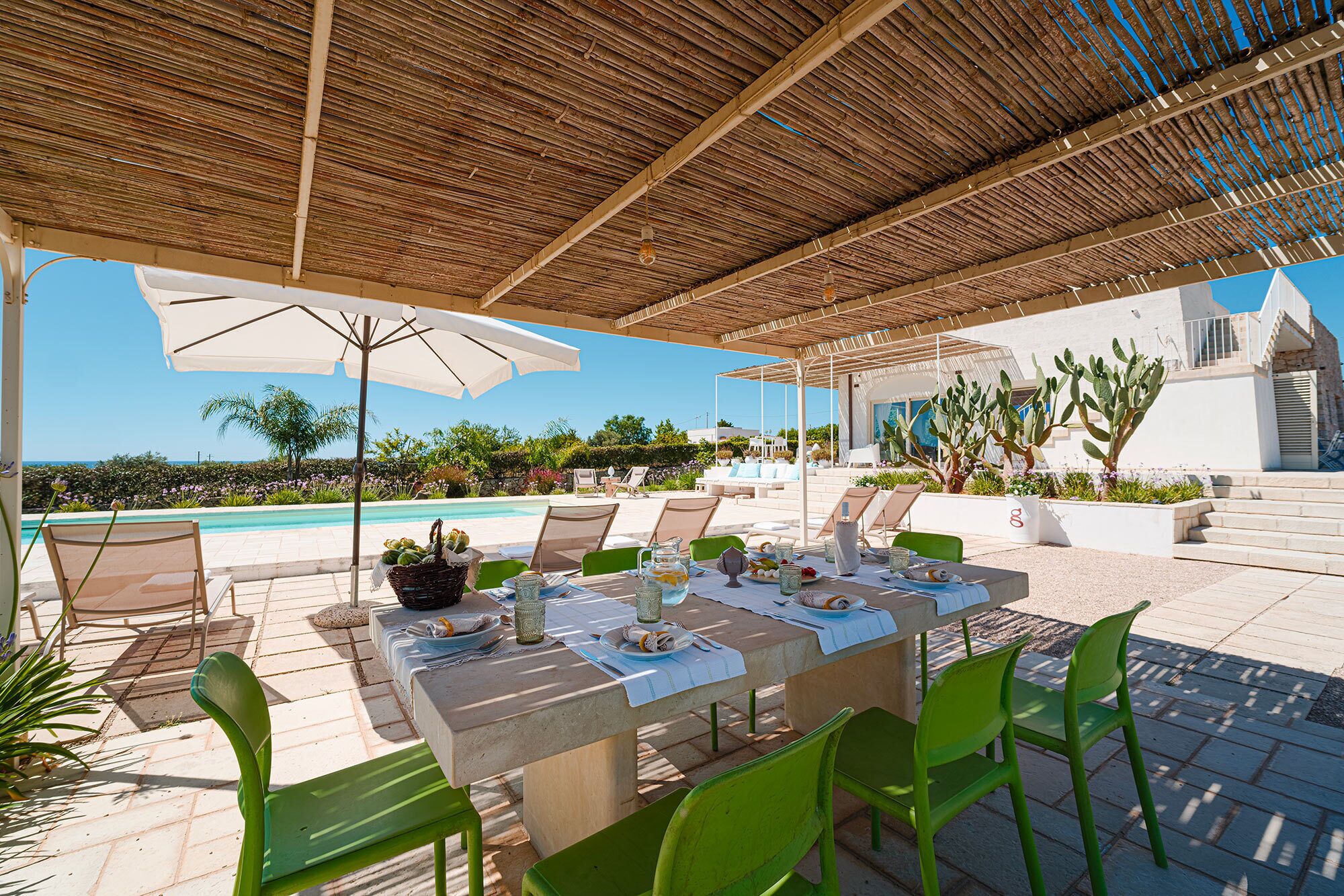 An outdoor dining area with a table set for four, surrounded by green chairs, under a wooden pergola with a view of the pool and landscape.