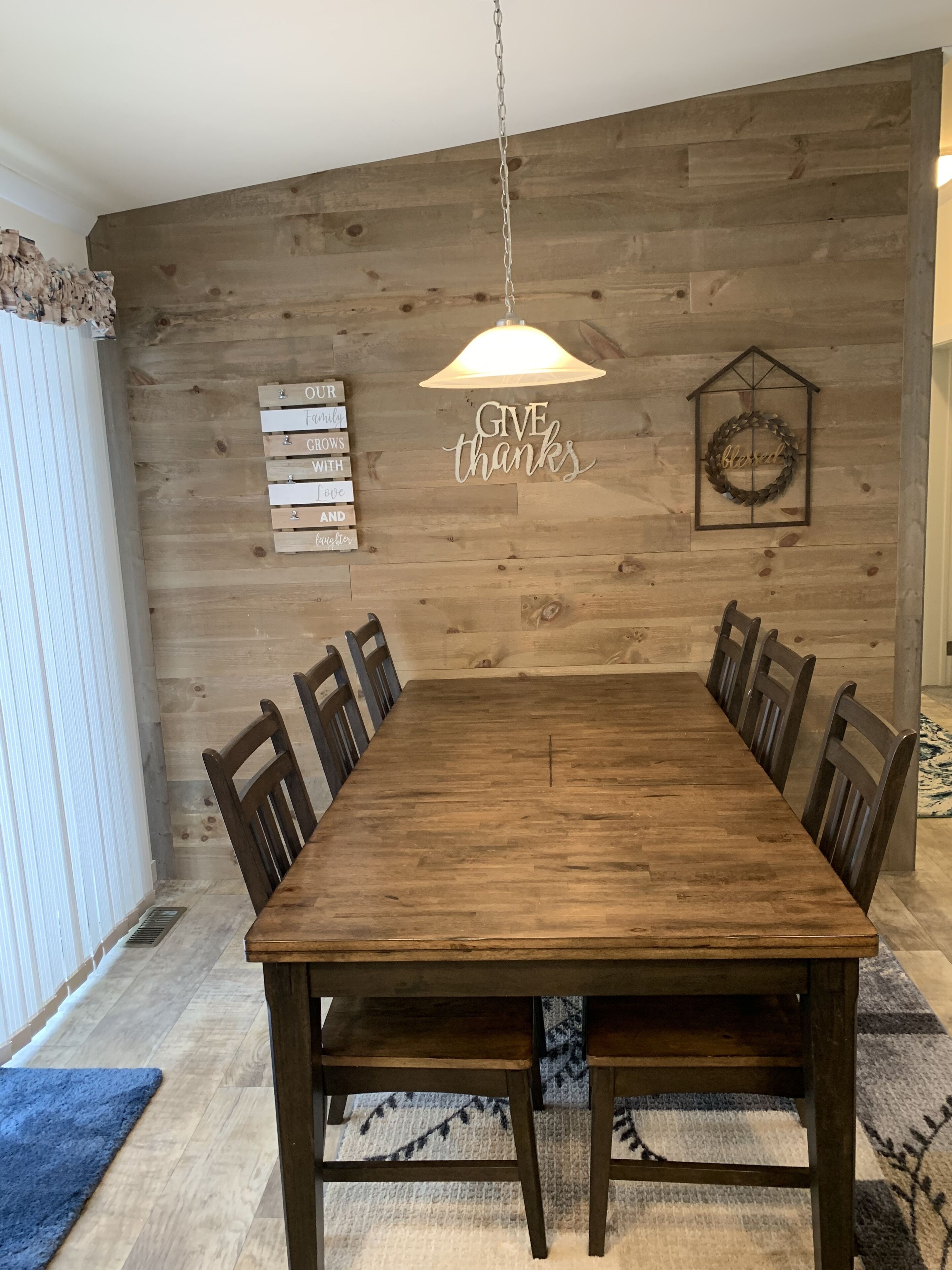 A dining room with a wooden table and chairs, a hanging light fixture, and a wall with a 'GIVE thanks' sign and a wreath.
