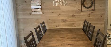 A dining room with a wooden table and chairs, a hanging light fixture, and a wall with a 'GIVE thanks' sign and a wreath.