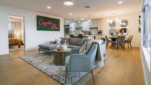 A modern living room with a sofa, armchair, and coffee table. There is a dining area with a table and chairs. A neon sign reads "Coyote Den." The room has wooden flooring and a patterned rug. - Luxury 5BR Old Town Villa with Heated Pool & Games (Scottsdale)