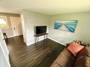 Relax in this cozy space with a flat-screen TV and a beautiful painting above, complemented by a comfortable couch and a stylish door leading to another room.
