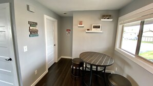 A room with a round table and two stools, a window with a view of a driveway, and various wall decorations including a sign that says "Cabin Rules Be Ready & Go" and "Camp Fires Eat & Drink Plenty Take a Nap Enjoy the day". - Unit B Minimalist Living, Simply...Perfect! (Kalispell)