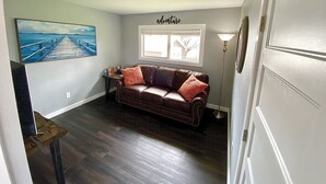 A living room with a brown leather sofa, a painting of a pier, a window with a view, and a lamp. - Unit B Minimalist Living, Simply...Perfect! (Kalispell)