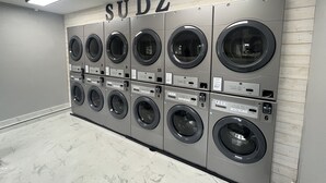 A row of six wall-mounted washing machines in a laundry room with a sign above them reading "SUDZ". - Unit B Minimalist Living, Simply...Perfect! (Kalispell)