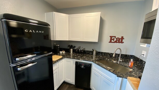 A compact kitchen with white cabinets, a black refrigerator, a granite countertop, and a stainless steel sink. - Unit B Minimalist Living, Simply...Perfect! (Kalispell)