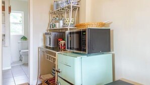 Cocina compacta con refrigerador antiguo, microondas y horno pequeño. Hay un armario de madera a la derecha y una puerta que conduce a otra habitación.