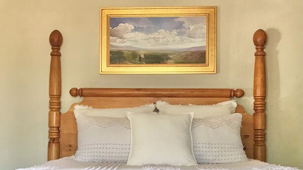 Una cama con cabecera de madera, ropa de cama blanca y un cuadro de paisaje enmarcado sobre ella.