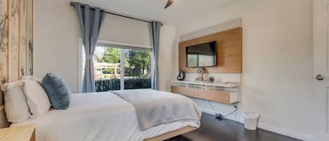 Quarto com cama, ventilador de teto, TV fixada na parede e janela com cortinas.